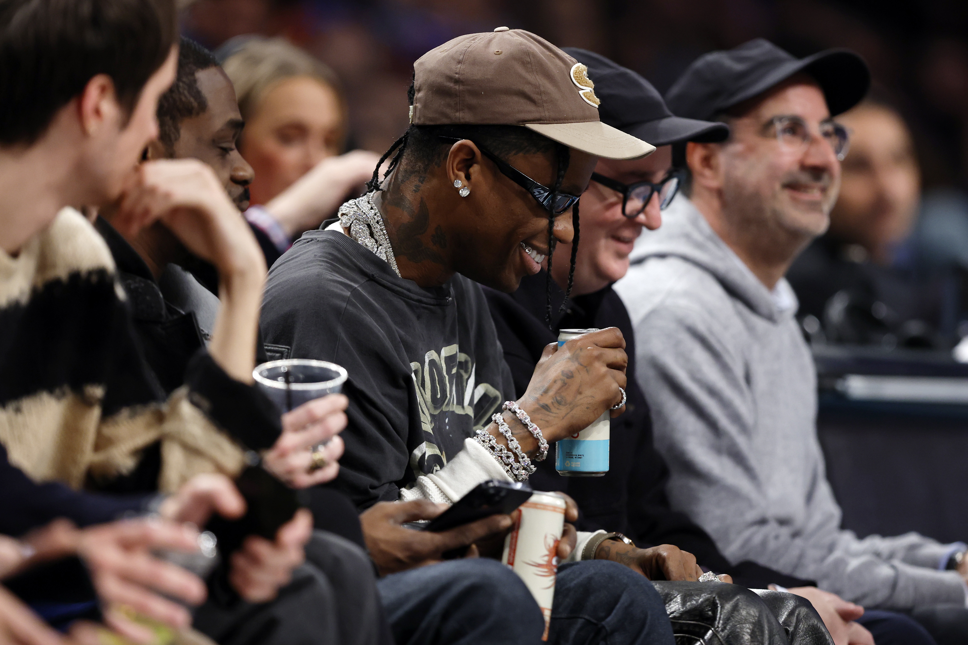 Travis Scott sitting courtside at a basketball game while holding a can of his Cacti drink
