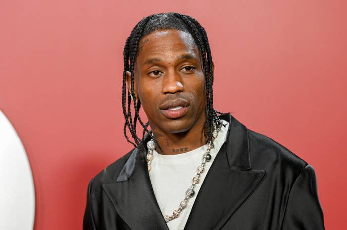 Travis in a shirt and jacket with braided hair and sparkling necklace, looking off-camera