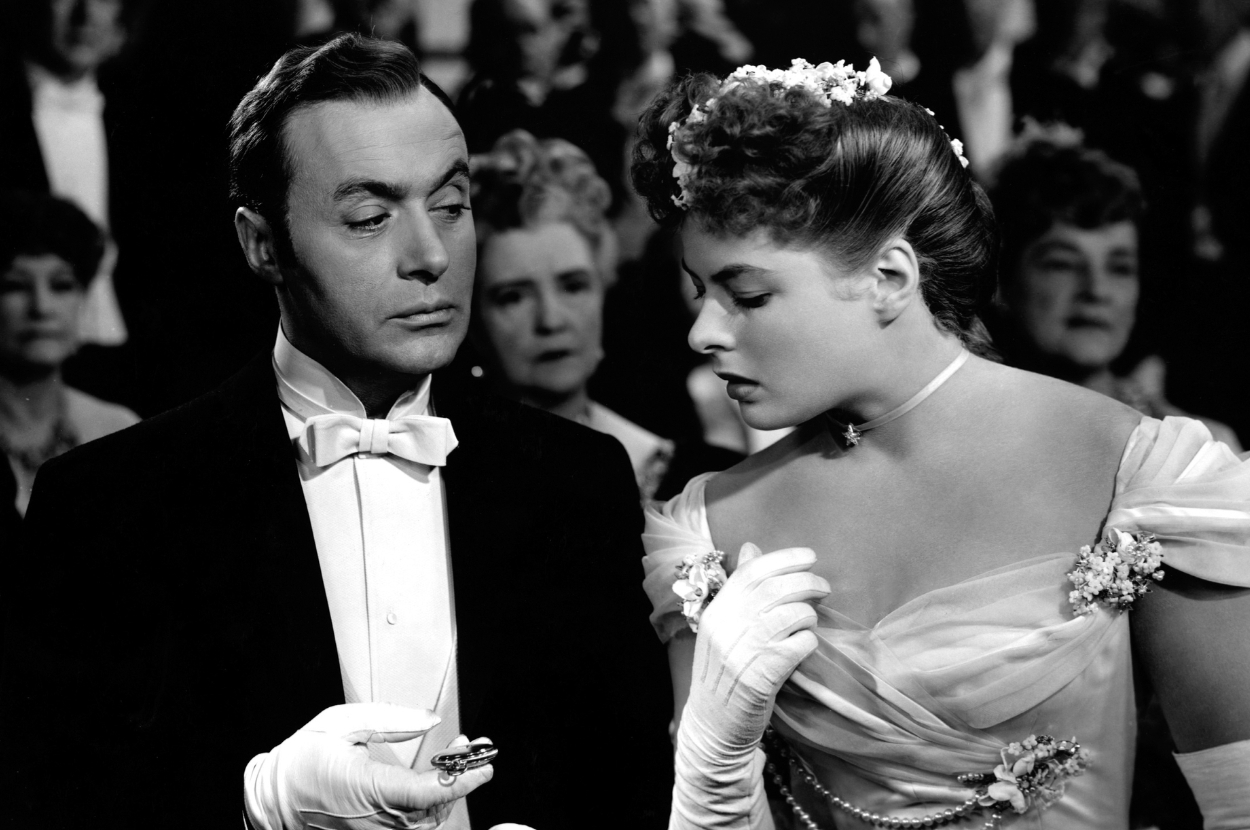 Man in suit with bow tie and woman in off-shoulder dress with flower in hair; both looking at a small object in man&#x27;s hand