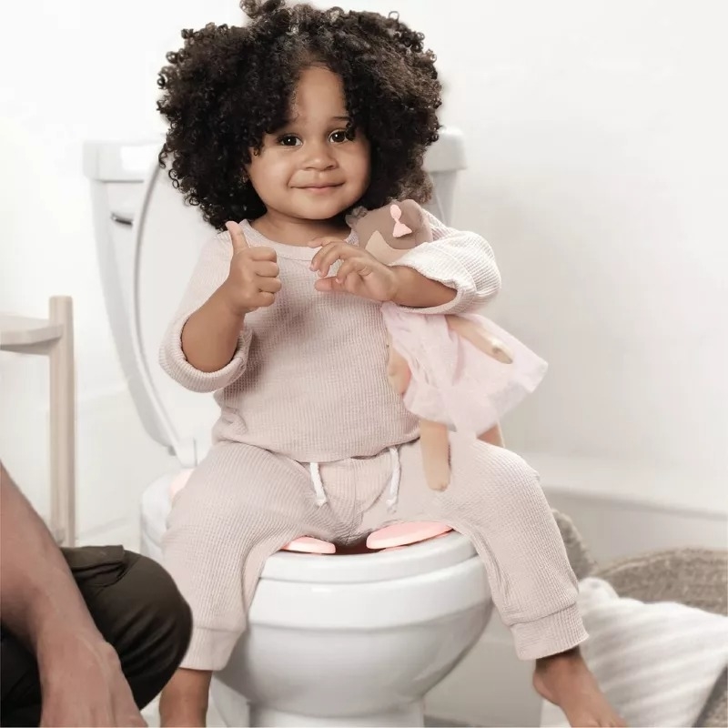 Child on a potty training seat, holding a doll, giving a thumbs up, with an adult&#x27;s leg nearby