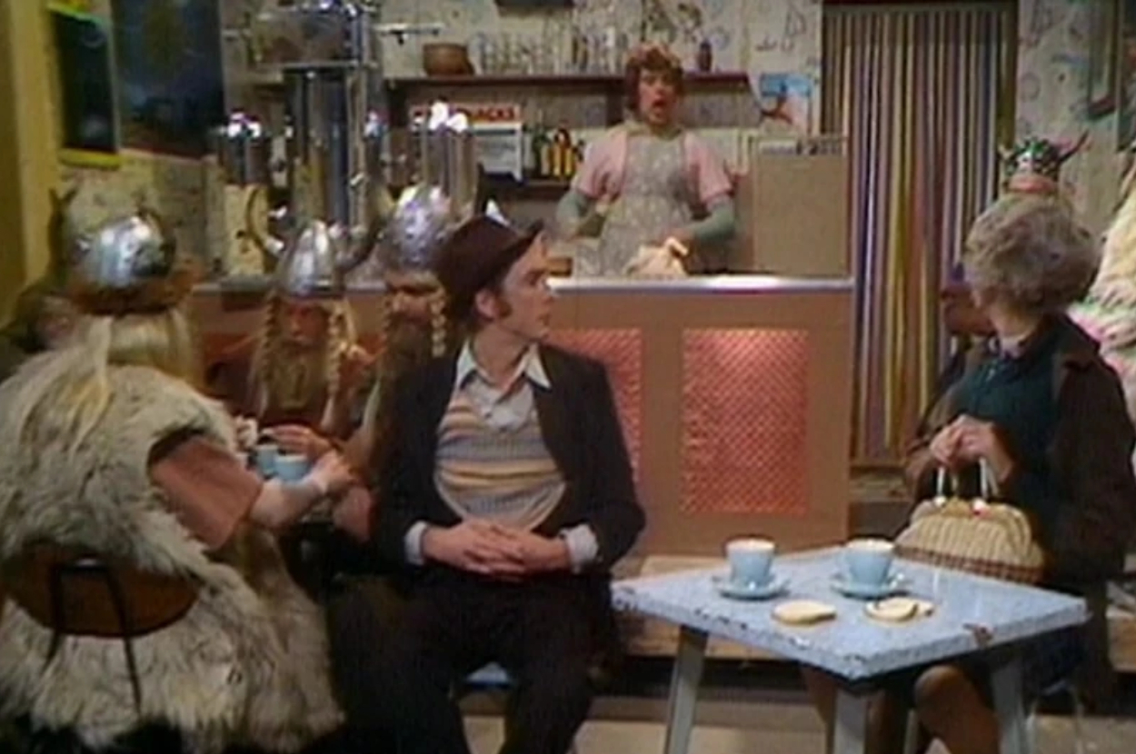 Scene from Monty Python&#x27;s Flying Circus with characters in a cafe, some wearing Viking helmets