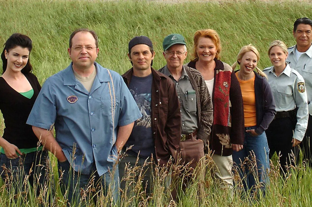 Group of 8 people standing in a field for a cast photo from the TV show &quot;Corner Gas.&quot;
