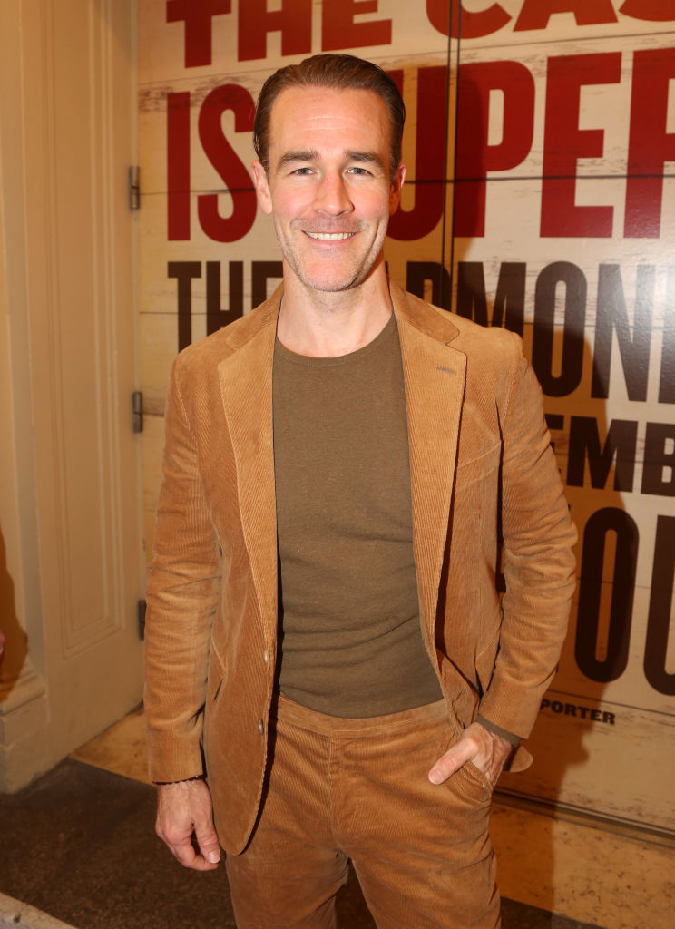 James standing, smiling in a corduroy suit without a tie, hands in pockets, against a backdrop with text