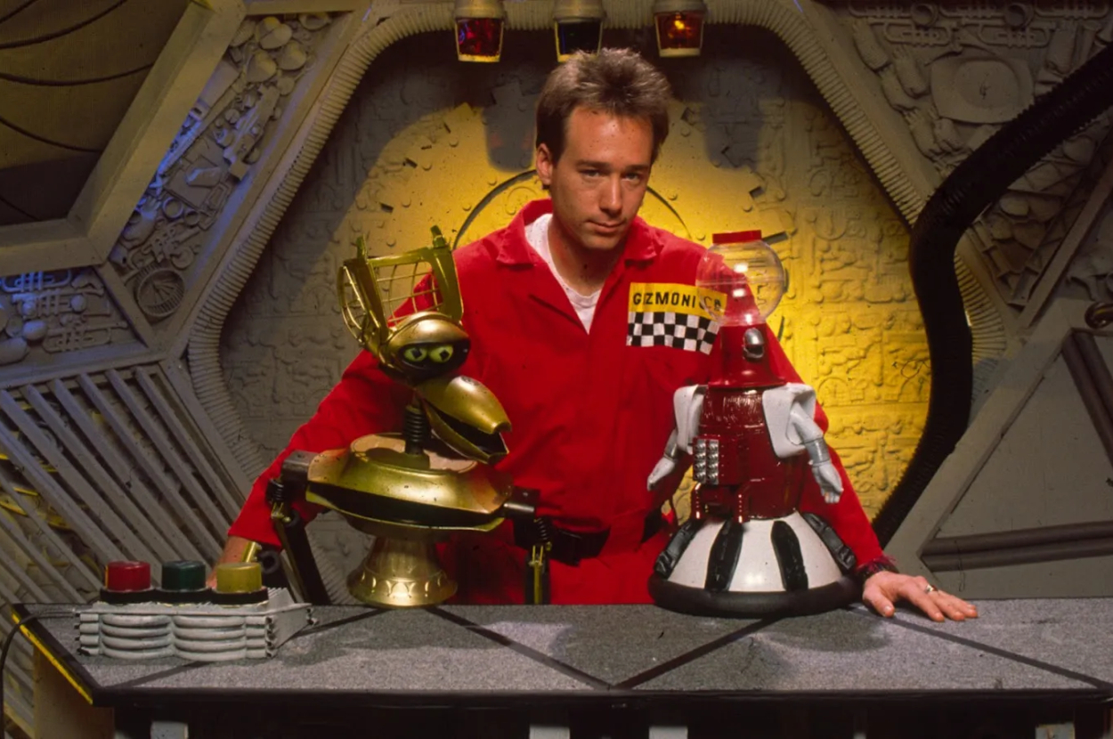 Man in red jumpsuit with two robot puppets in a spaceship set