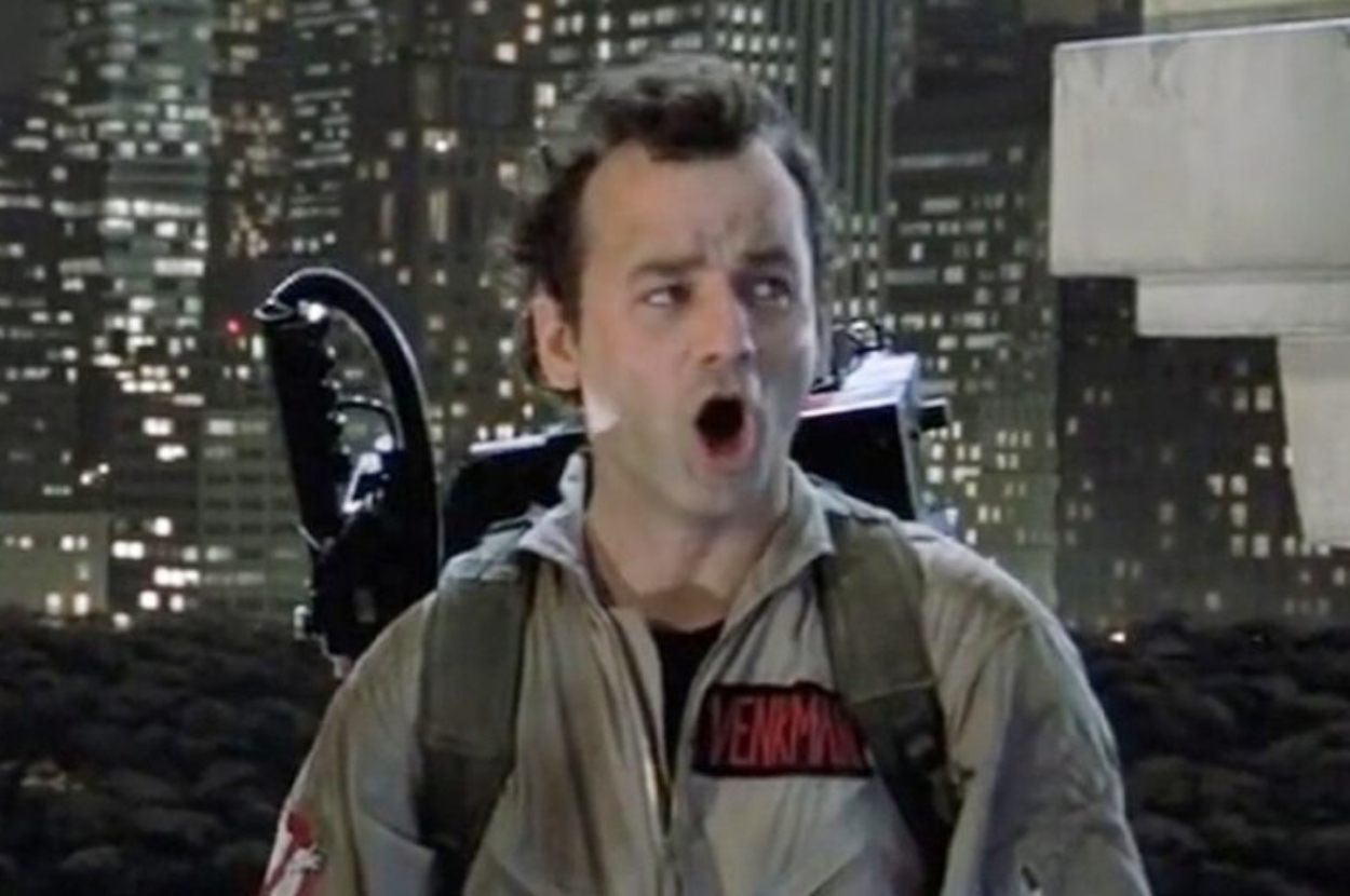 Bill Murray in a Ghostbusters uniform, looking surprised, with New York City night skyline in the background