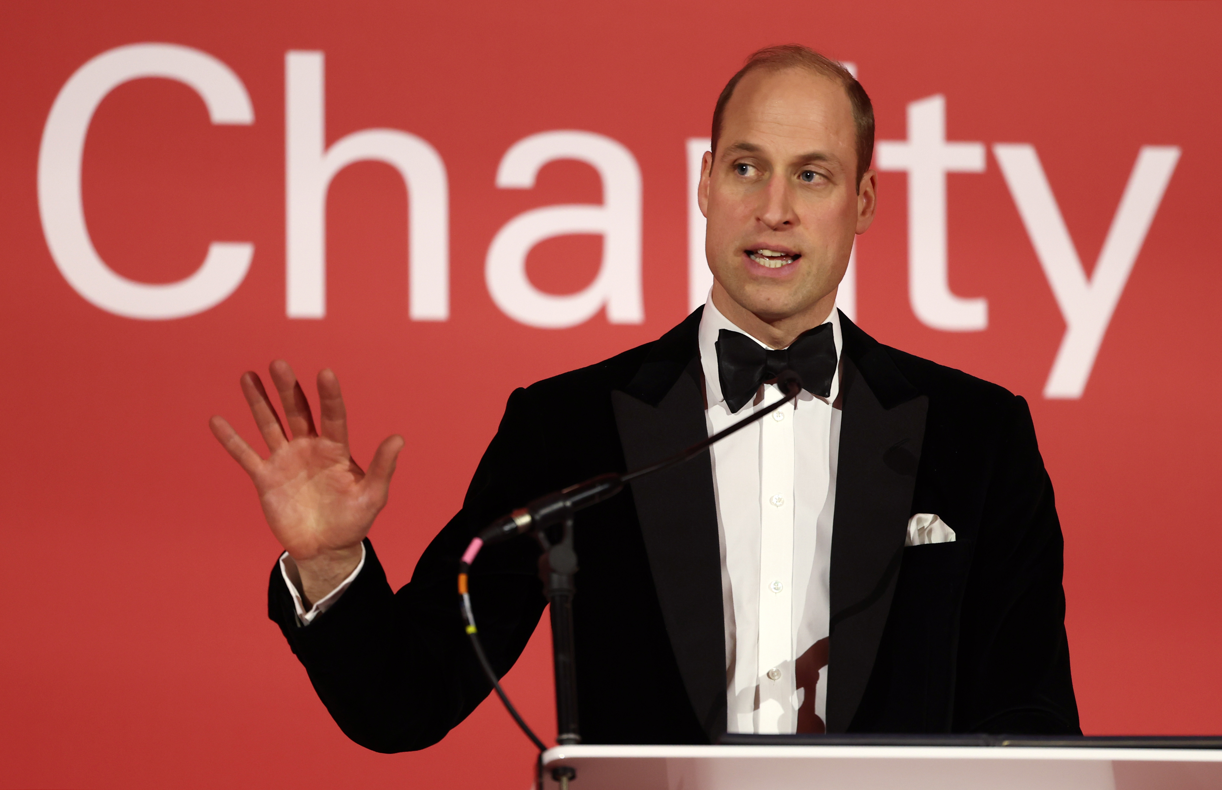 William in a tuxedo with a bow tie speaks at a podium with a &quot;Charity&quot; sign in the background