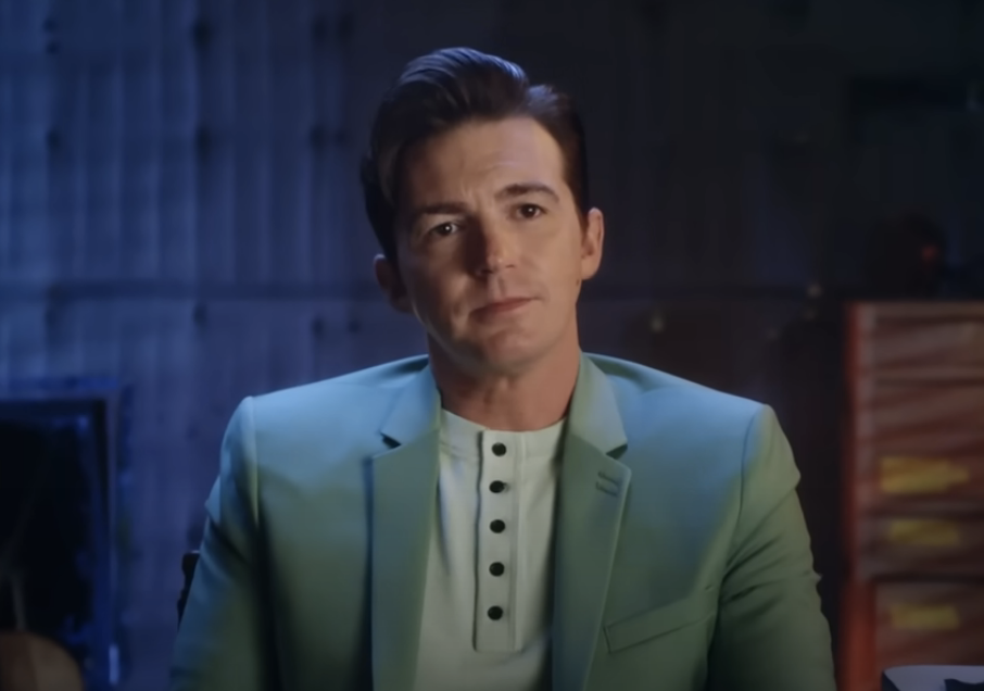Drake Bell in a light suit on set, with a serious expression