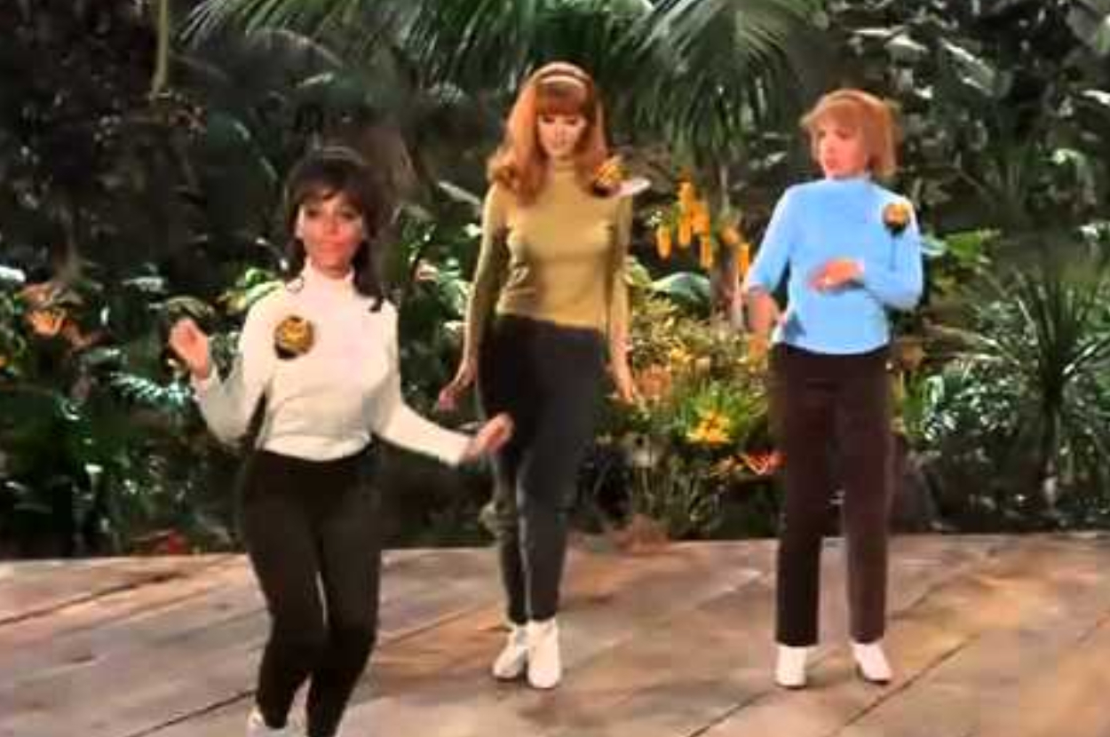 Three women from the TV show &#x27;Gilligan&#x27;s Island&#x27; stand together, wearing casual 60s outfits with themed patches