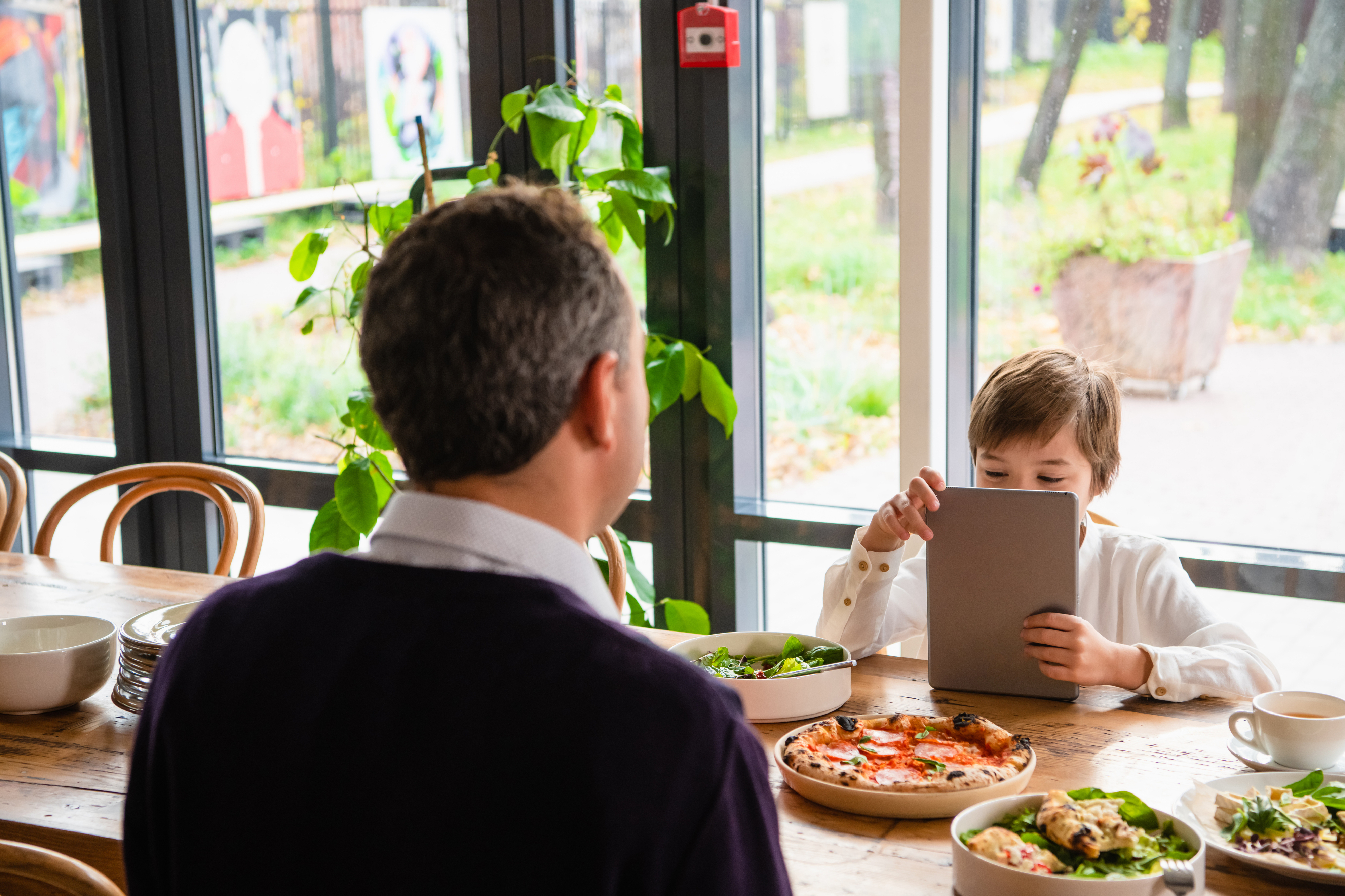 Two individuals at a restaurant table, one using a tablet, with pizza and salads in front of them