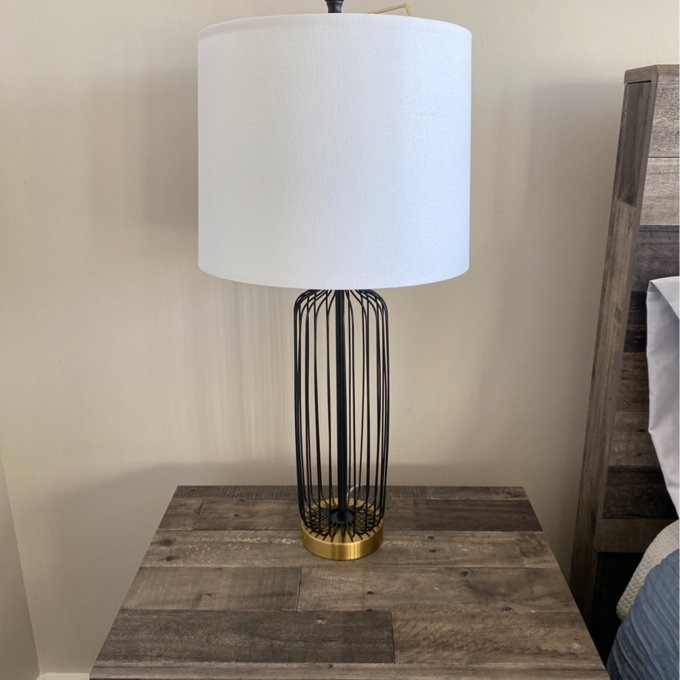 Table lamp with cylindrical open framework base and a white shade, on a wood nightstand. Suitable for home decor