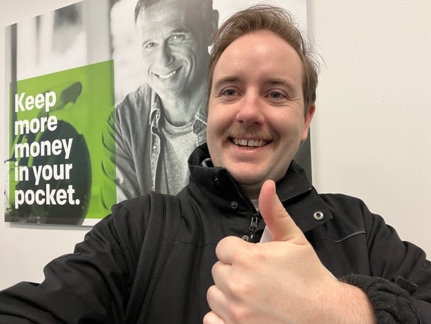 Man smiling with thumbs up, standing in front of a poster with another smiling man and text about saving money