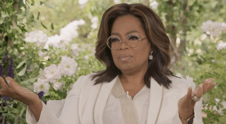 Oprah Winfrey, seated, gesturing, wearing glasses and a white outfit with earrings