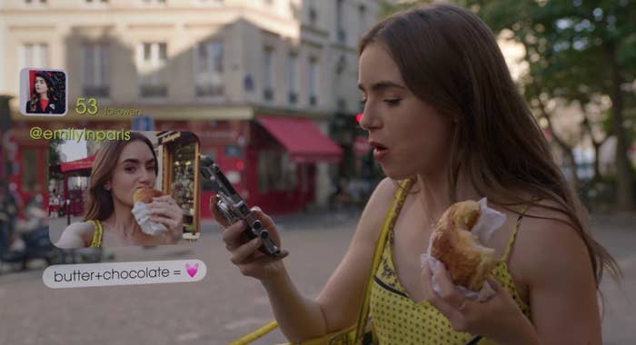 Woman in a yellow dress takes a selfie while eating a croissant, with phone displaying a social media app