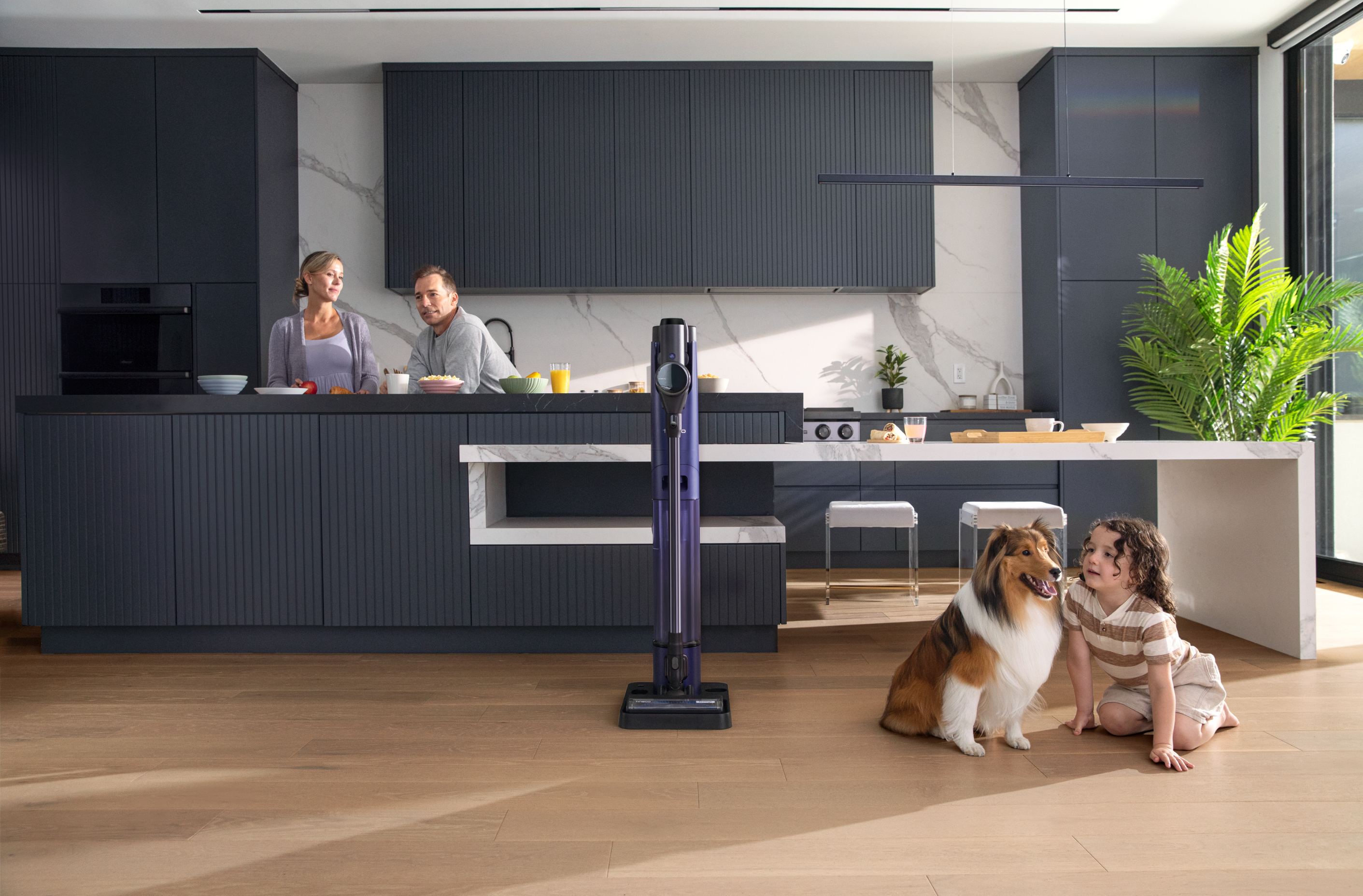 Family and dog in kitchen, child plays with vacuum, adults in background preparing food