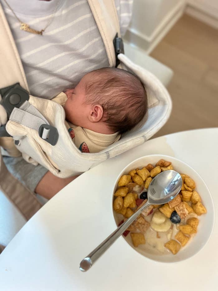 Adult holding a sleeping baby in a carrier beside a bowl of cereal with fruit