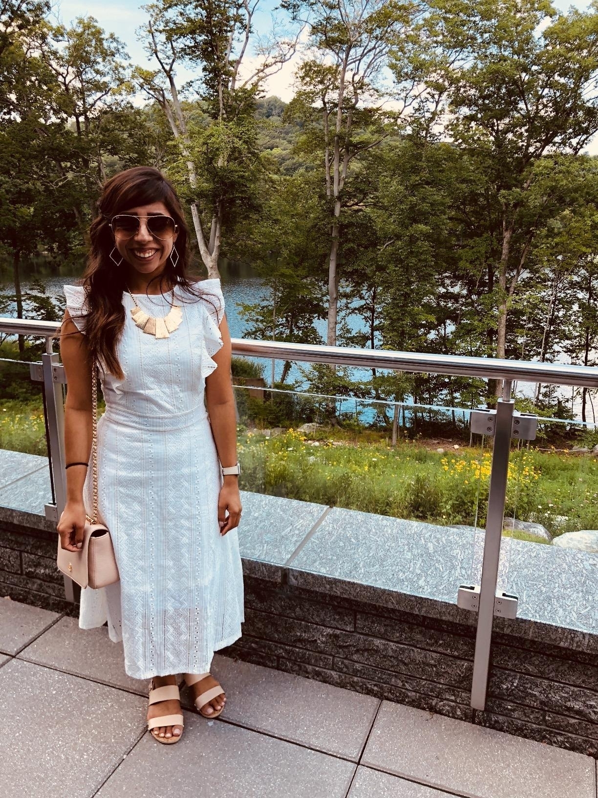 a reviewer in a sleeveless white dress with a necklace, sunglasses, and a handbag, standing by a railing with nature in the background