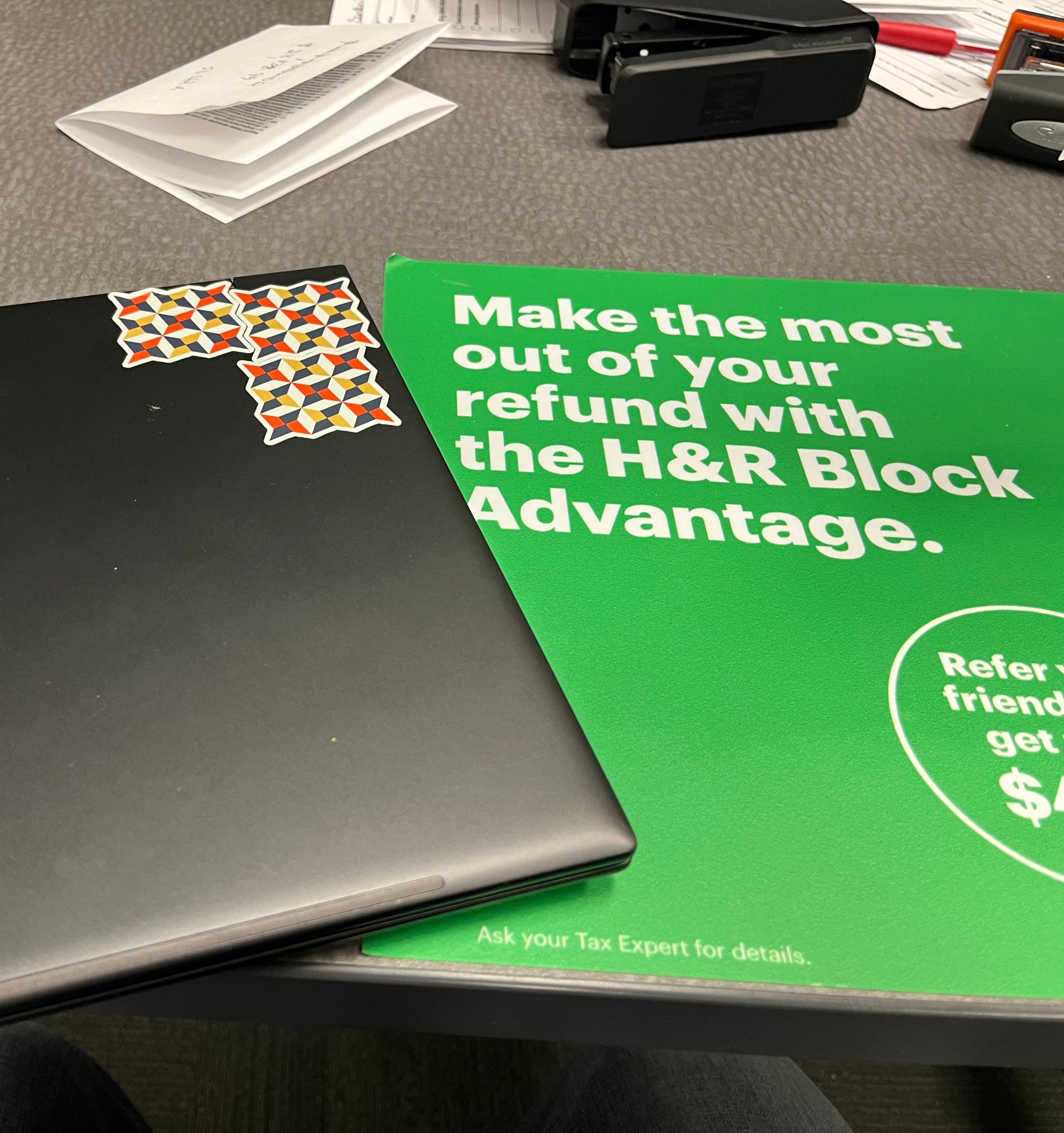 Un ordinateur portable avec des autocollants à côté d&#x27;une brochure verte de H&amp;amp;R Block sur un bureau avec des fournitures de bureau.