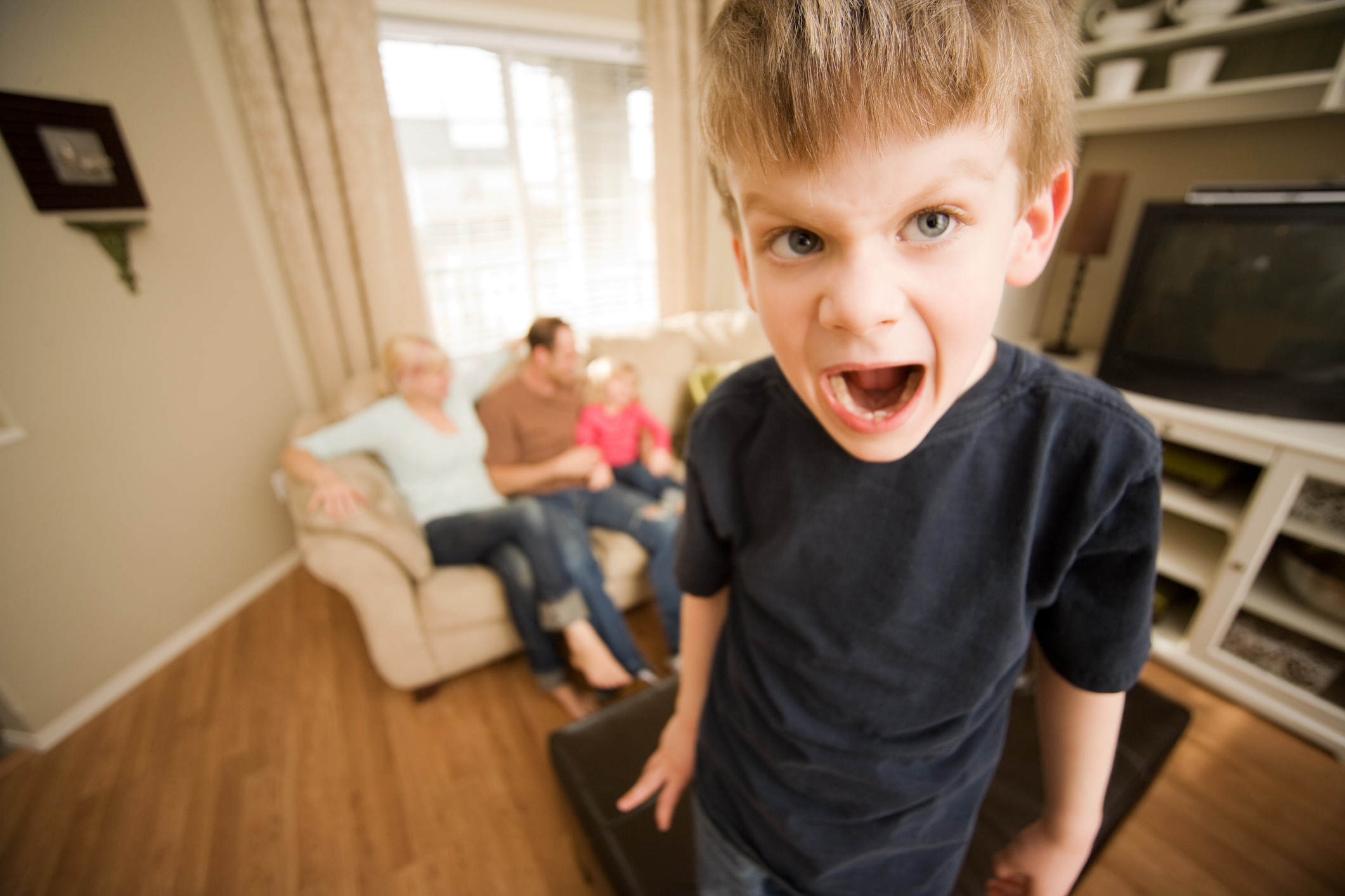 Young boy with mouth open in a playful roar, family in the background on a couch, indoors
