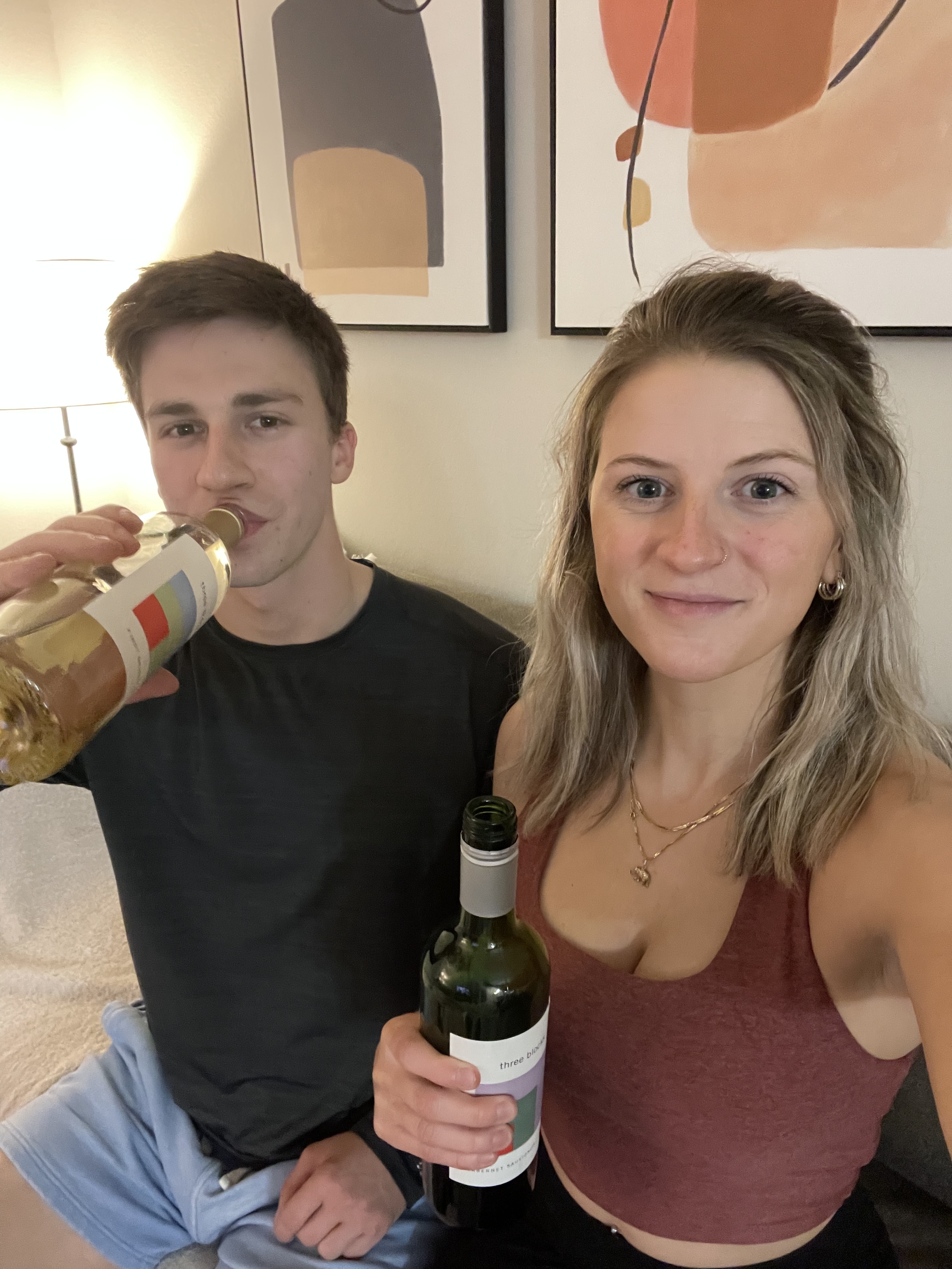 Man and woman smiling, holding wine bottles, casual attire indoors