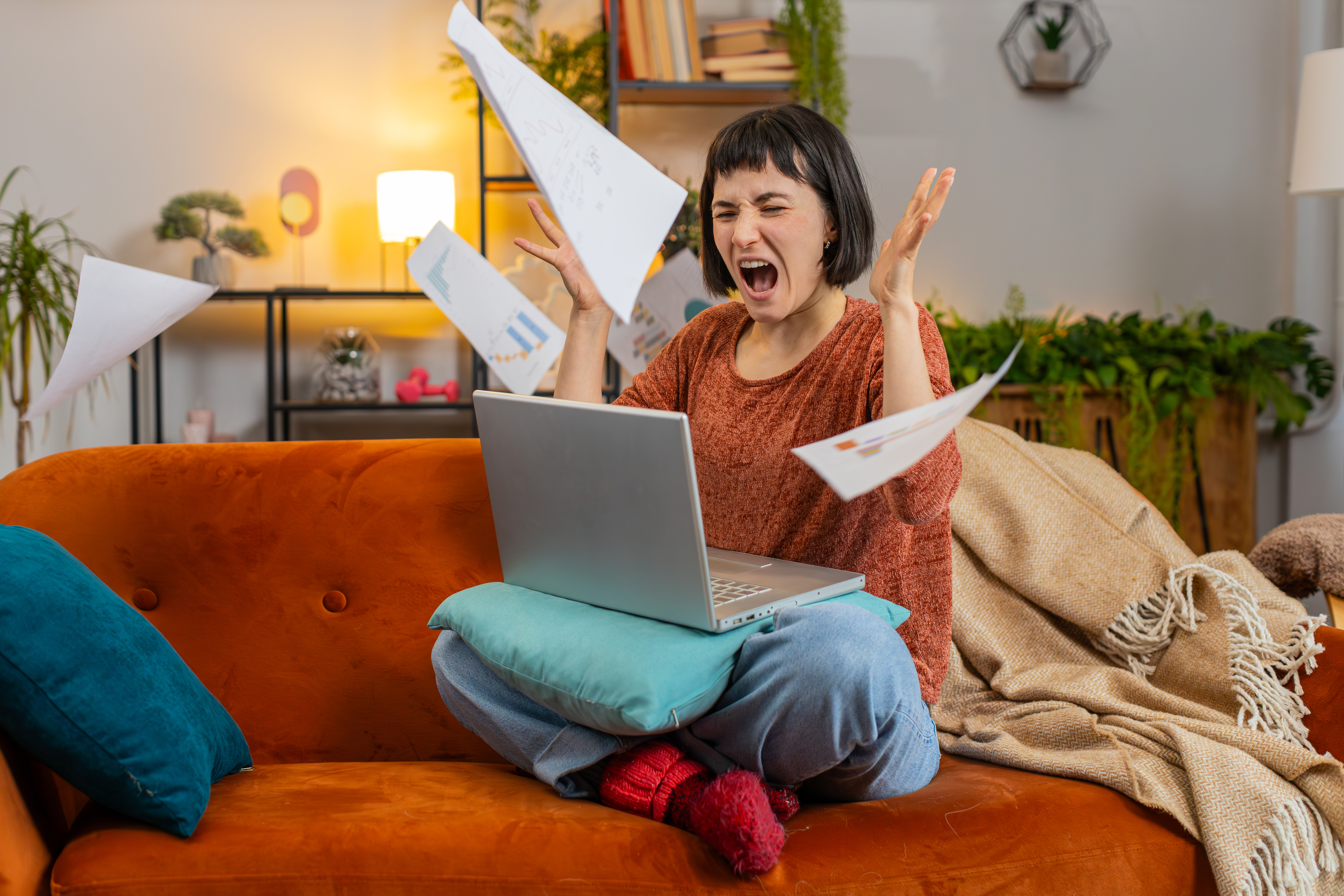 Woman sitting cross-legged on sofa with laptop, papers flying around, expressing frustration or excitement