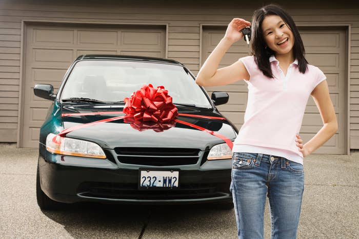 Young teenager standing next to a car with a large bow, smiling, hand on her head
