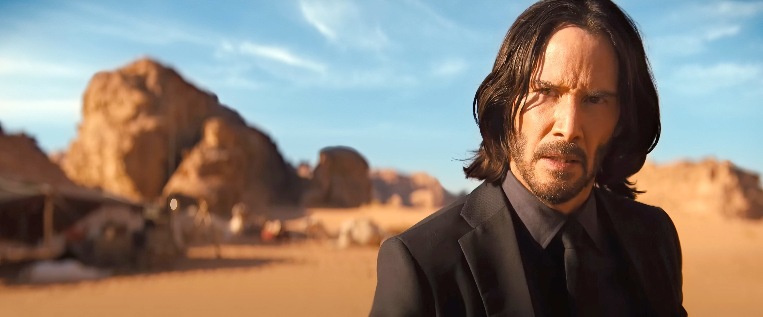 Keanu Reeves in a black suit standing in a desert with tents in the background