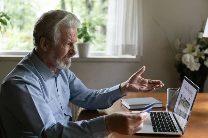 Older man gestures in frustration at laptop screen with visible video call, pen in hand, indoors