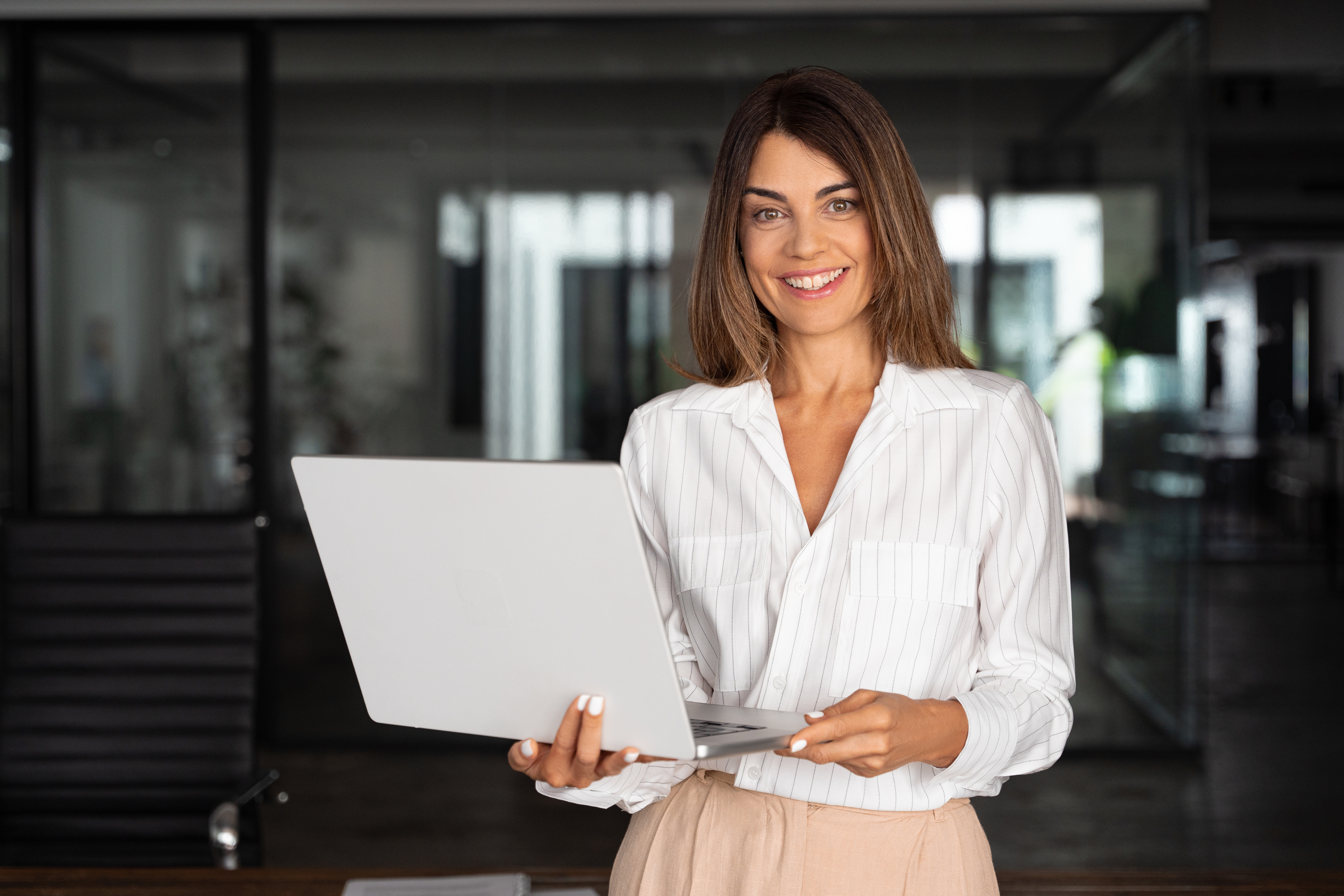 Woman holding a laptop in a business casual outfit, smiling at the camera