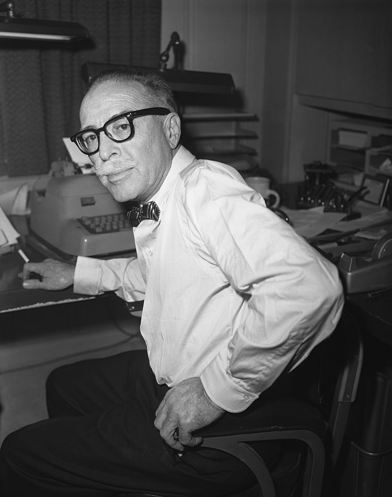 Dalton Trumbo in glasses and bow tie sitting at a desk with a typewriter, looking at the camera