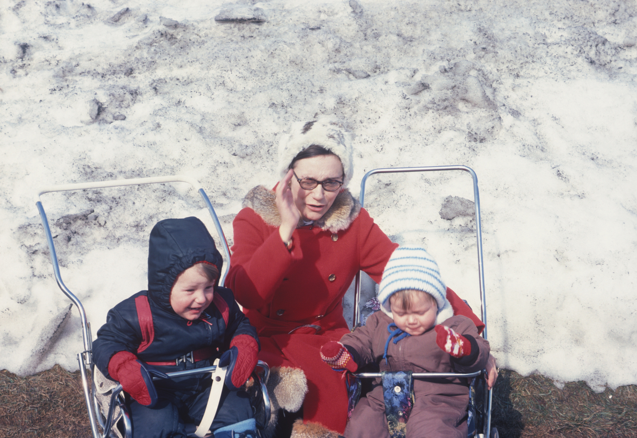 Woman seated between two children in sleds on snow, all wearing winter attire
