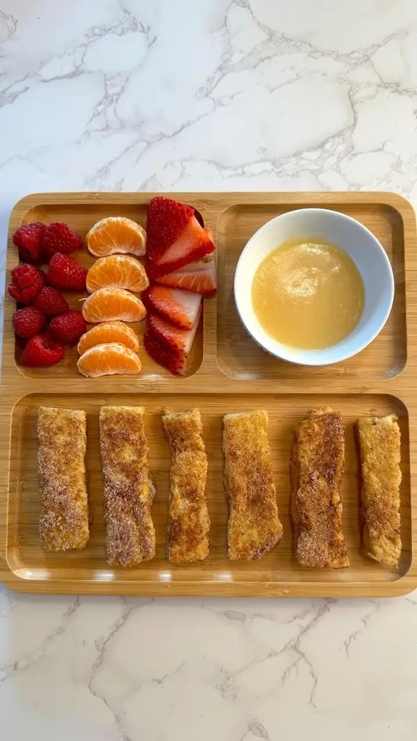 A plate with fruit slices, a bowl of dipping sauce, and breaded, fried rolls on a wooden tray