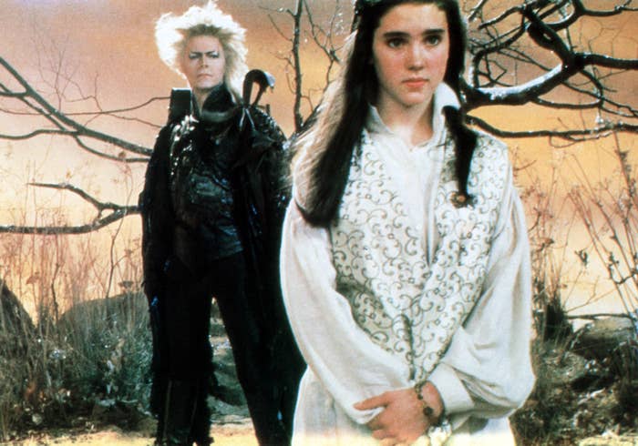 Jareth the Goblin King and Sarah stand in a mystical setting from the movie Labyrinth. Jareth is in a dark, embellished coat; Sarah wears a white dress
