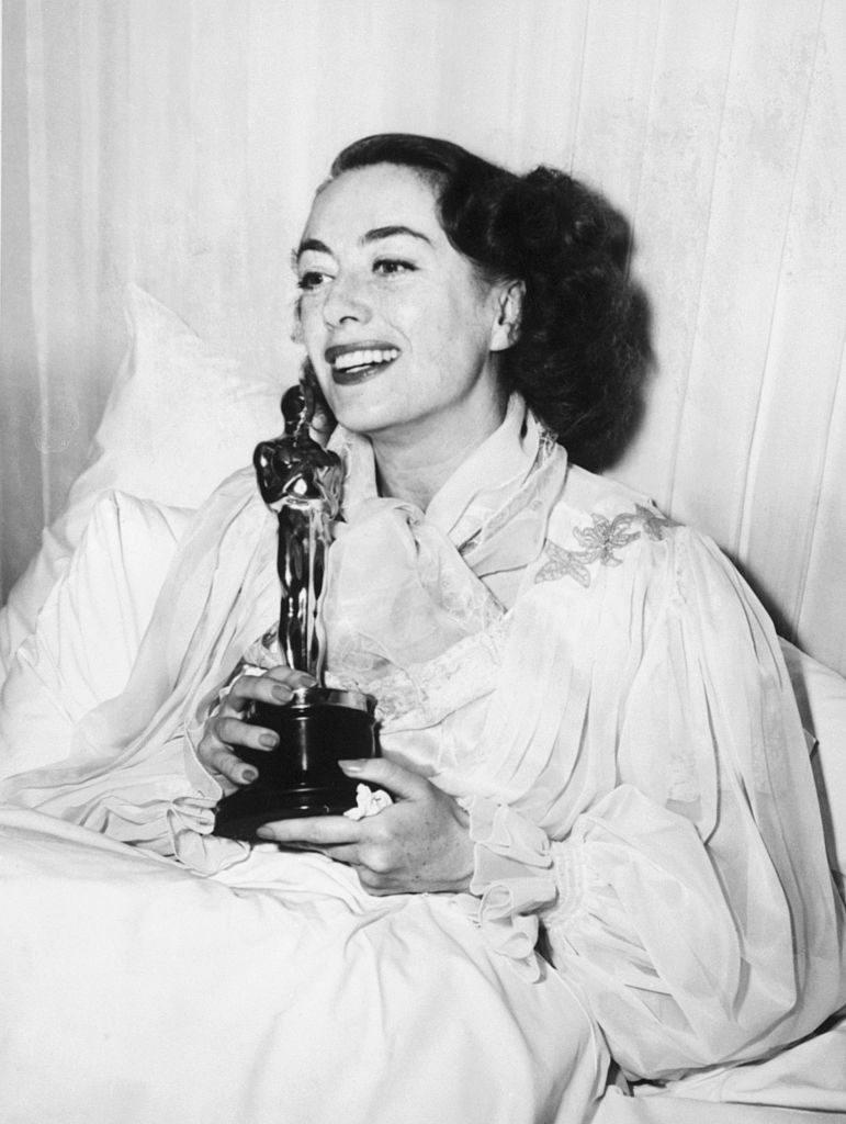 Joan in bed holding an Oscar trophy, wearing a ruffled gown with floral detail