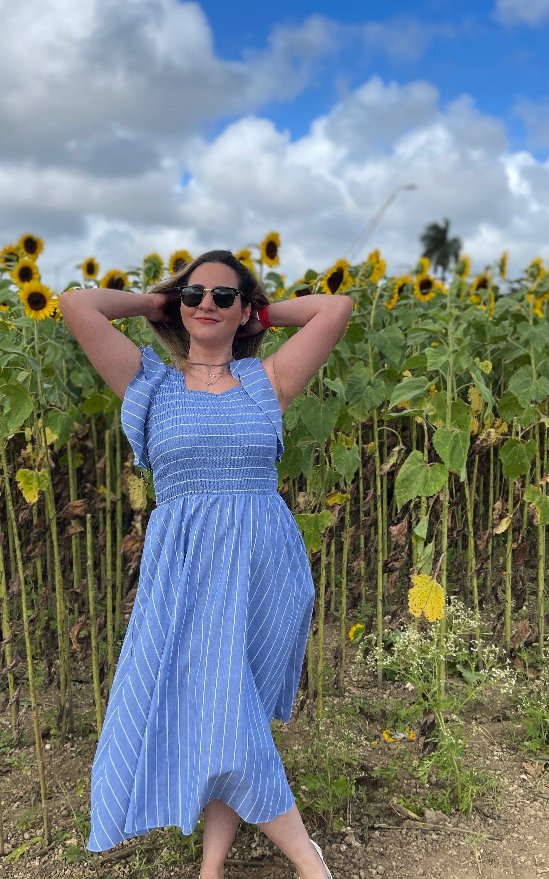 a reviewer in a blue striped sundress and sunglasses posing with hands behind head in a sunflower field