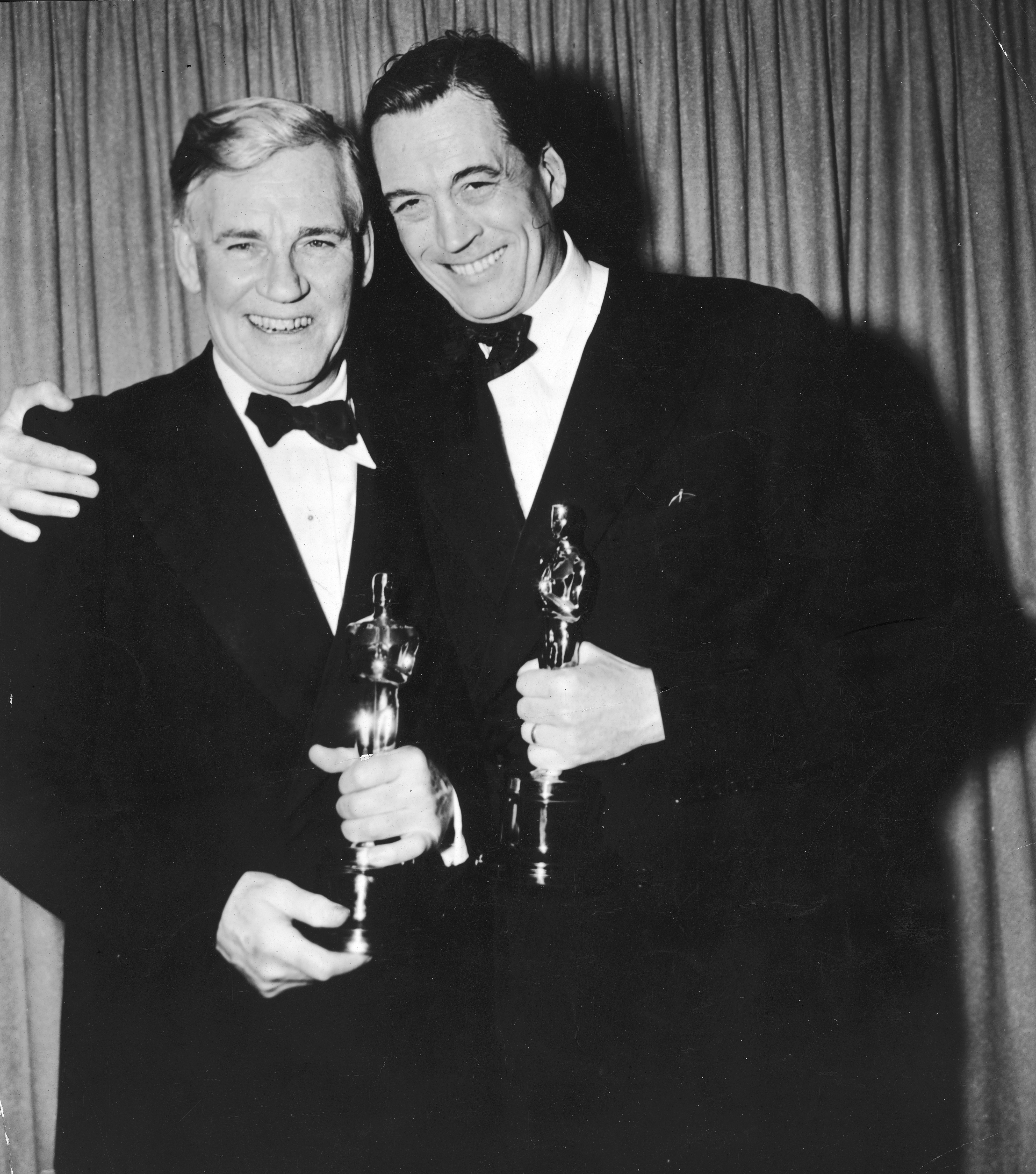 Two men in tuxedos holding award statues, smiling, with a curtain backdrop
