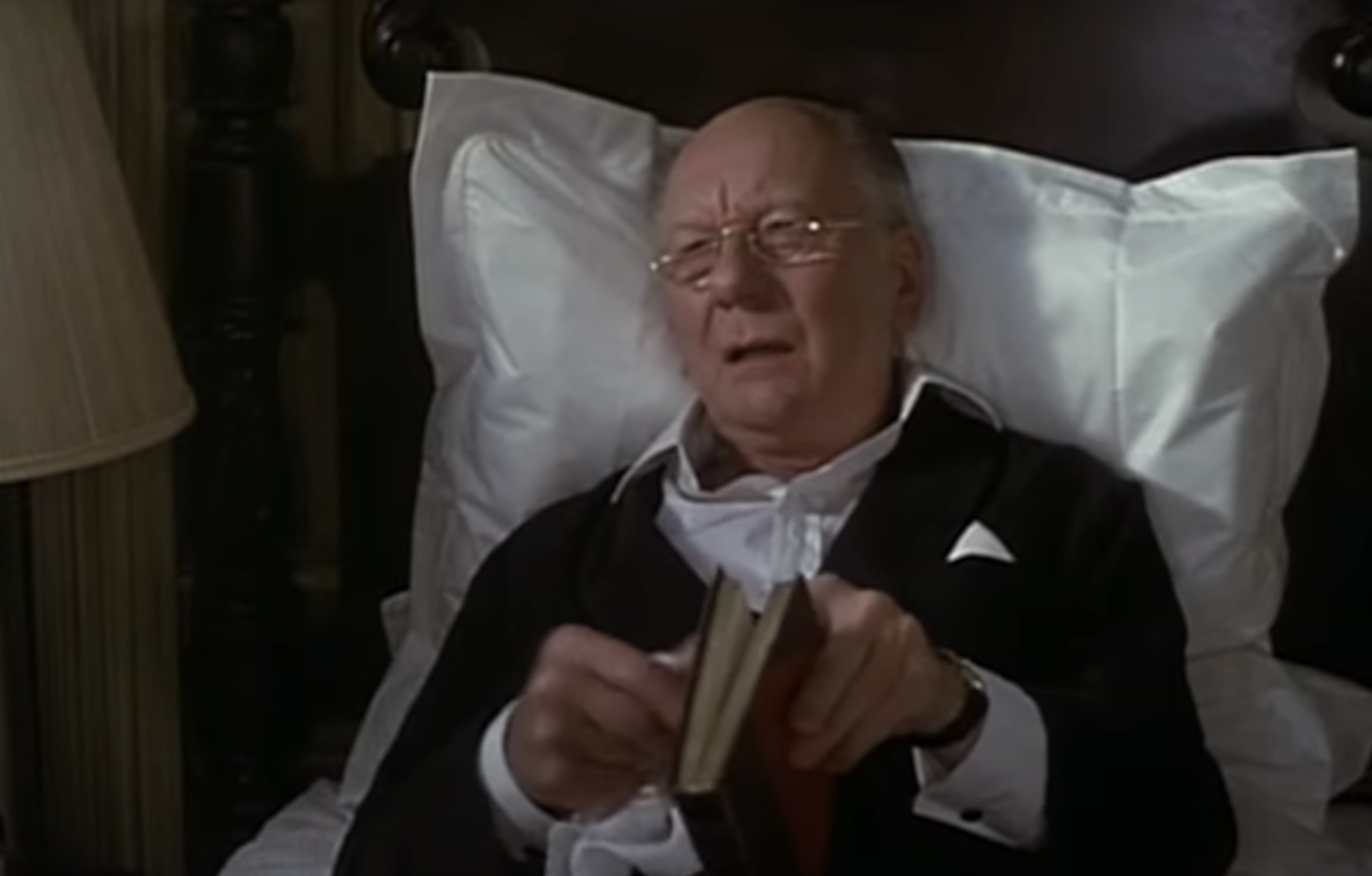 Elderly man in bed holding a book, looking pensive, from a film scene