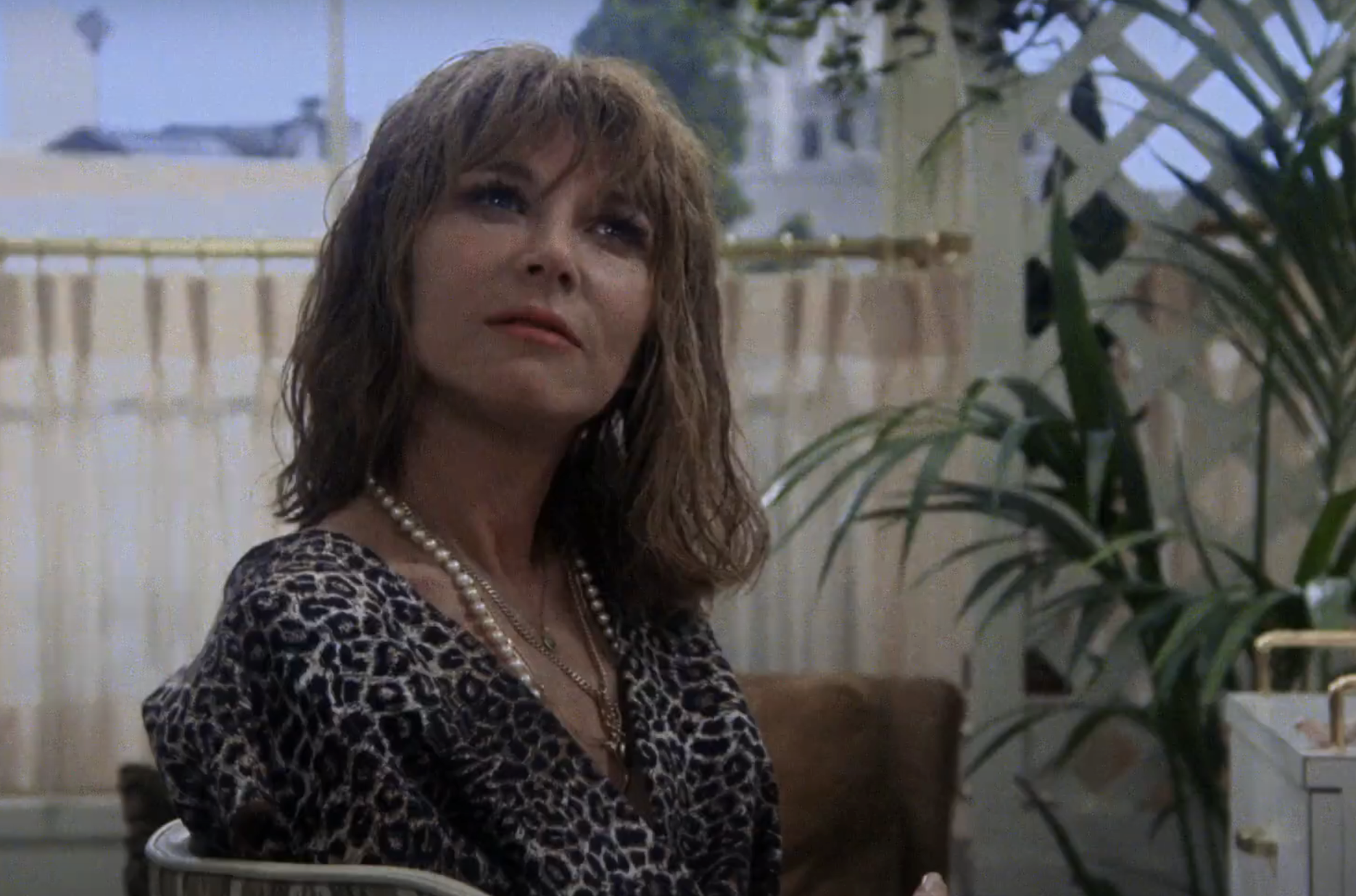 Lee Grant in a leopard print top and pearls, looking up, with potted plants in the background