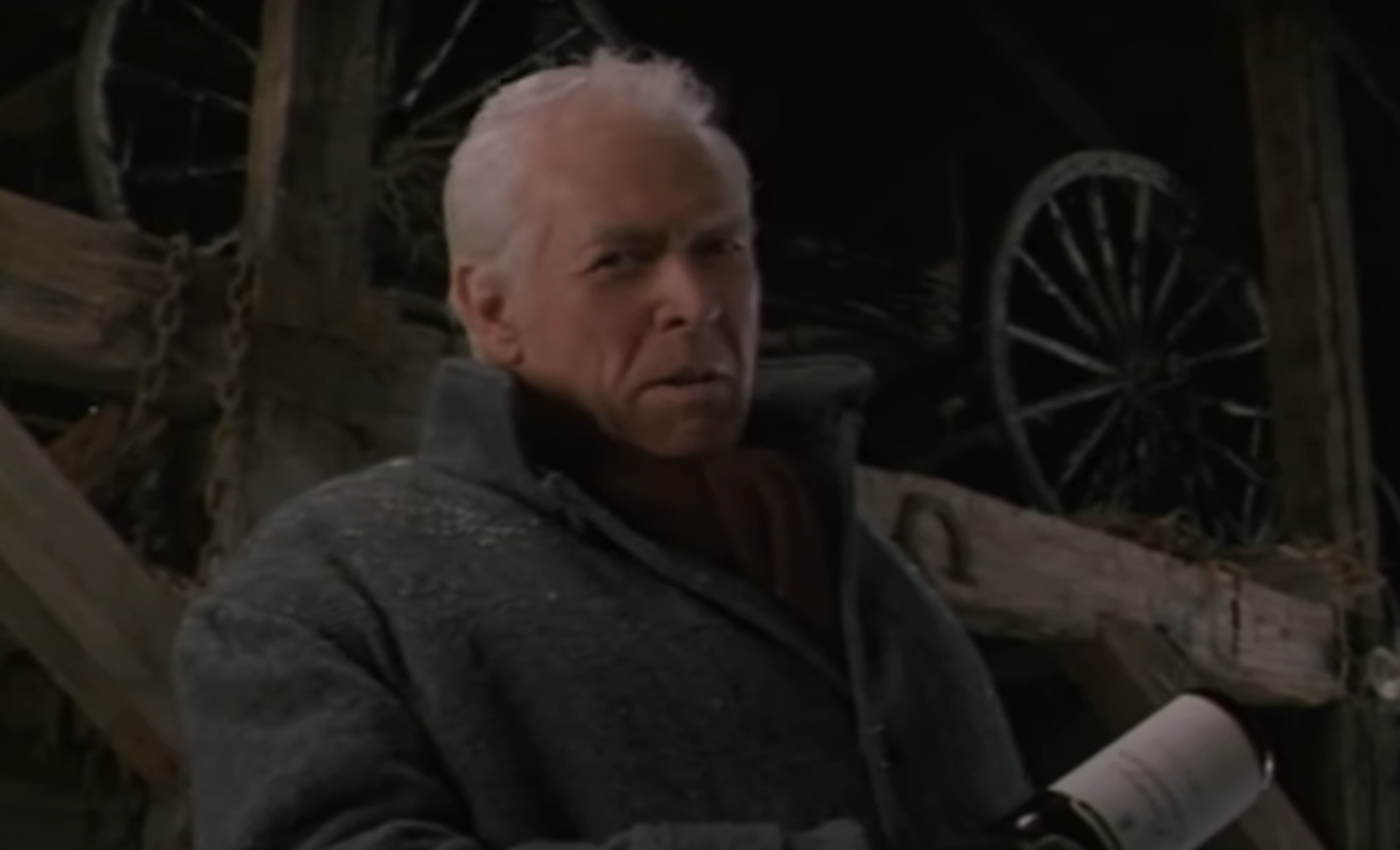 Clint Eastwood in a scene, wearing a coat, by a wooden structure