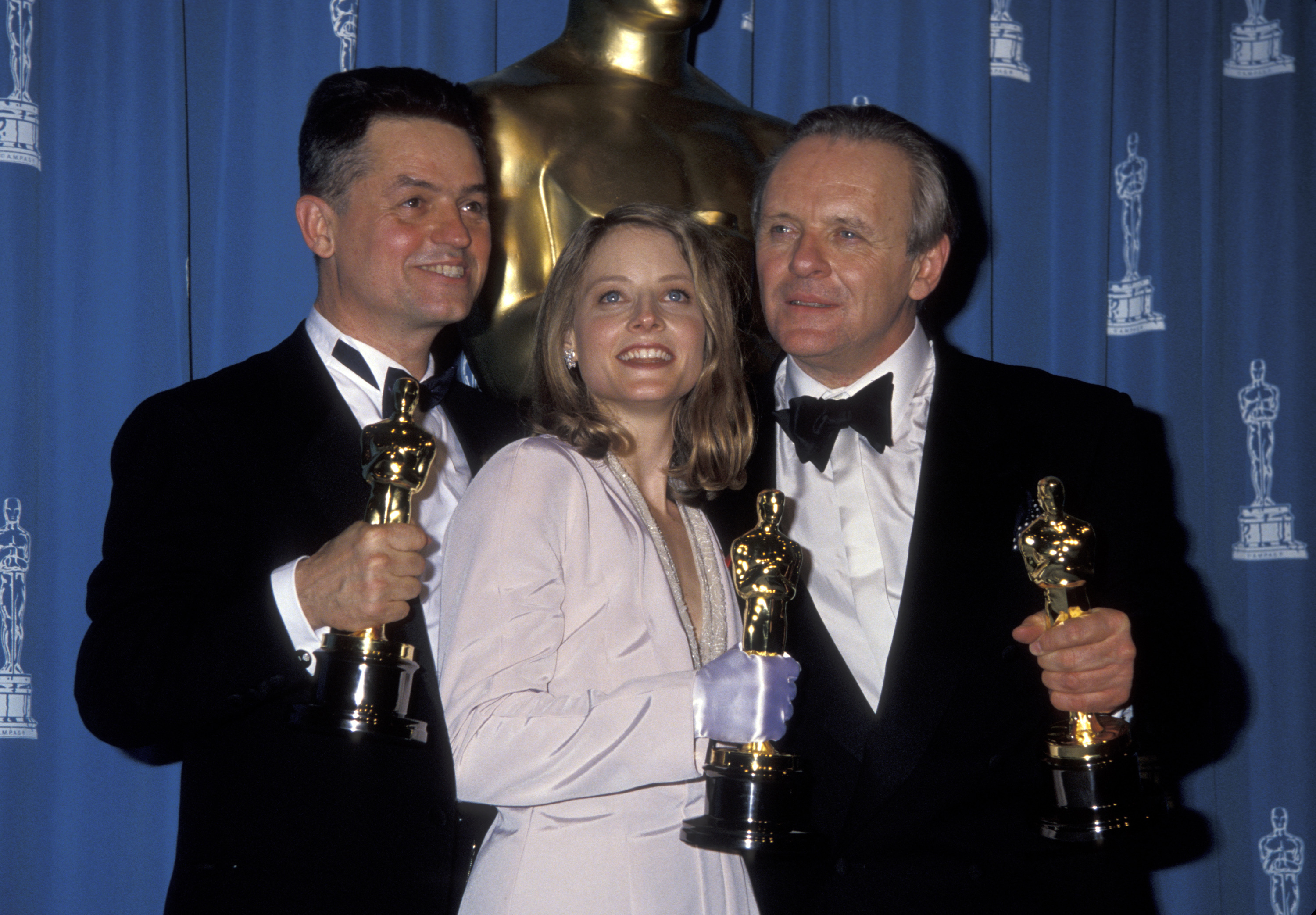 Three individuals at an awards ceremony holding trophies. Two in tuxedos, one in a blazer and dress