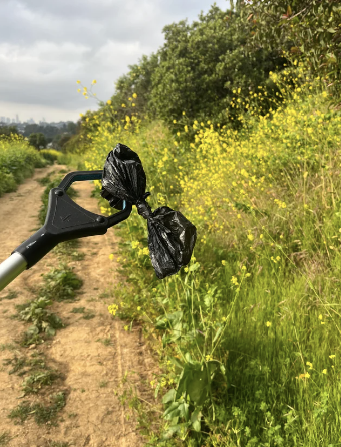 A litter grabber holds a tied black bag with the background of a nature trail and wildflowers