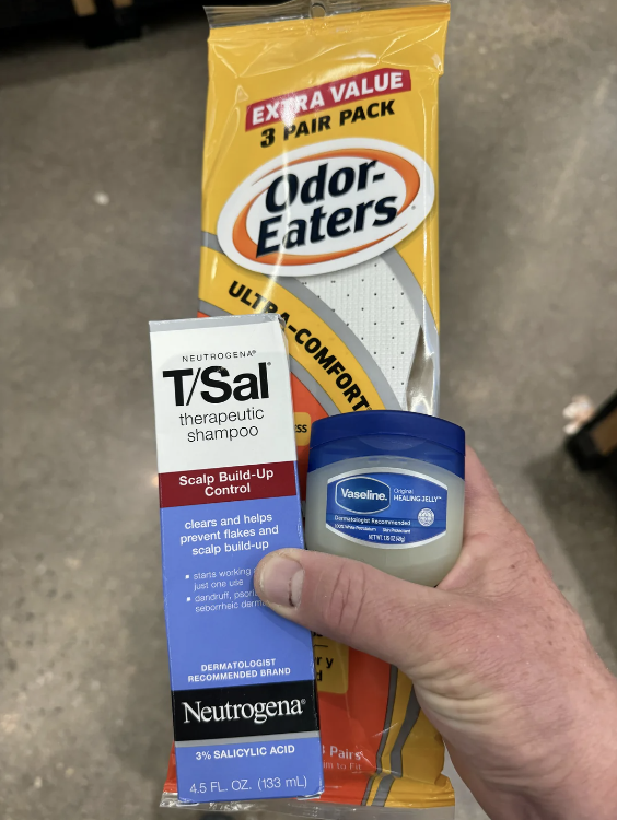 A hand holds a bottle of Neutrogena T/Sal shampoo next to a container of Vaseline and a pack of Odor-Eaters insoles on a store shelf