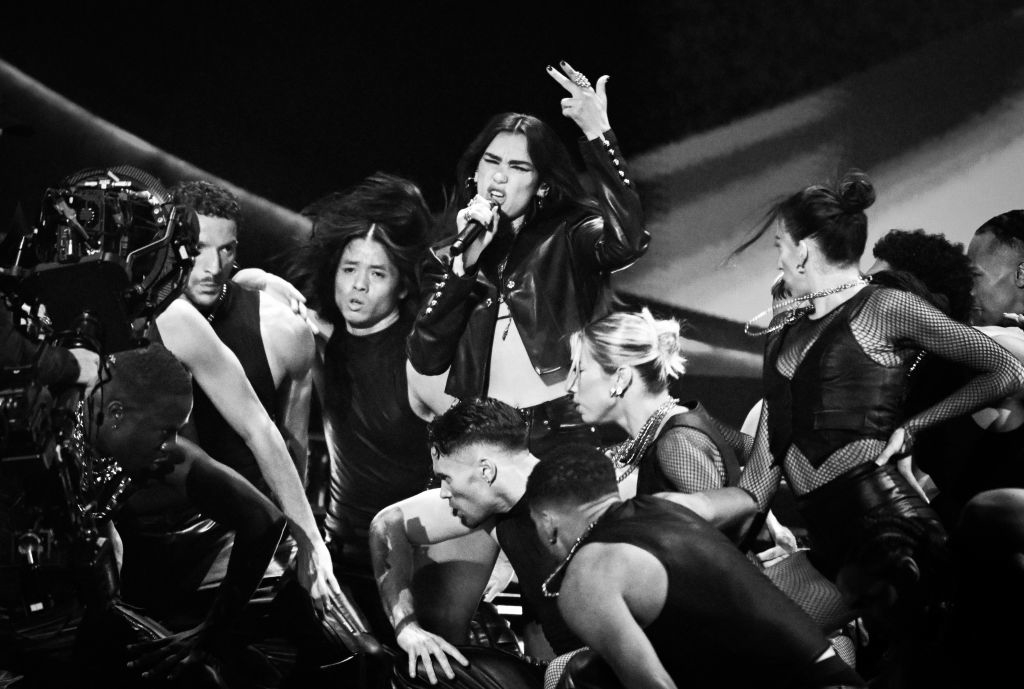 Performer singing on stage with a microphone, surrounded by dancers in coordinated black outfits. Camera equipment visible
