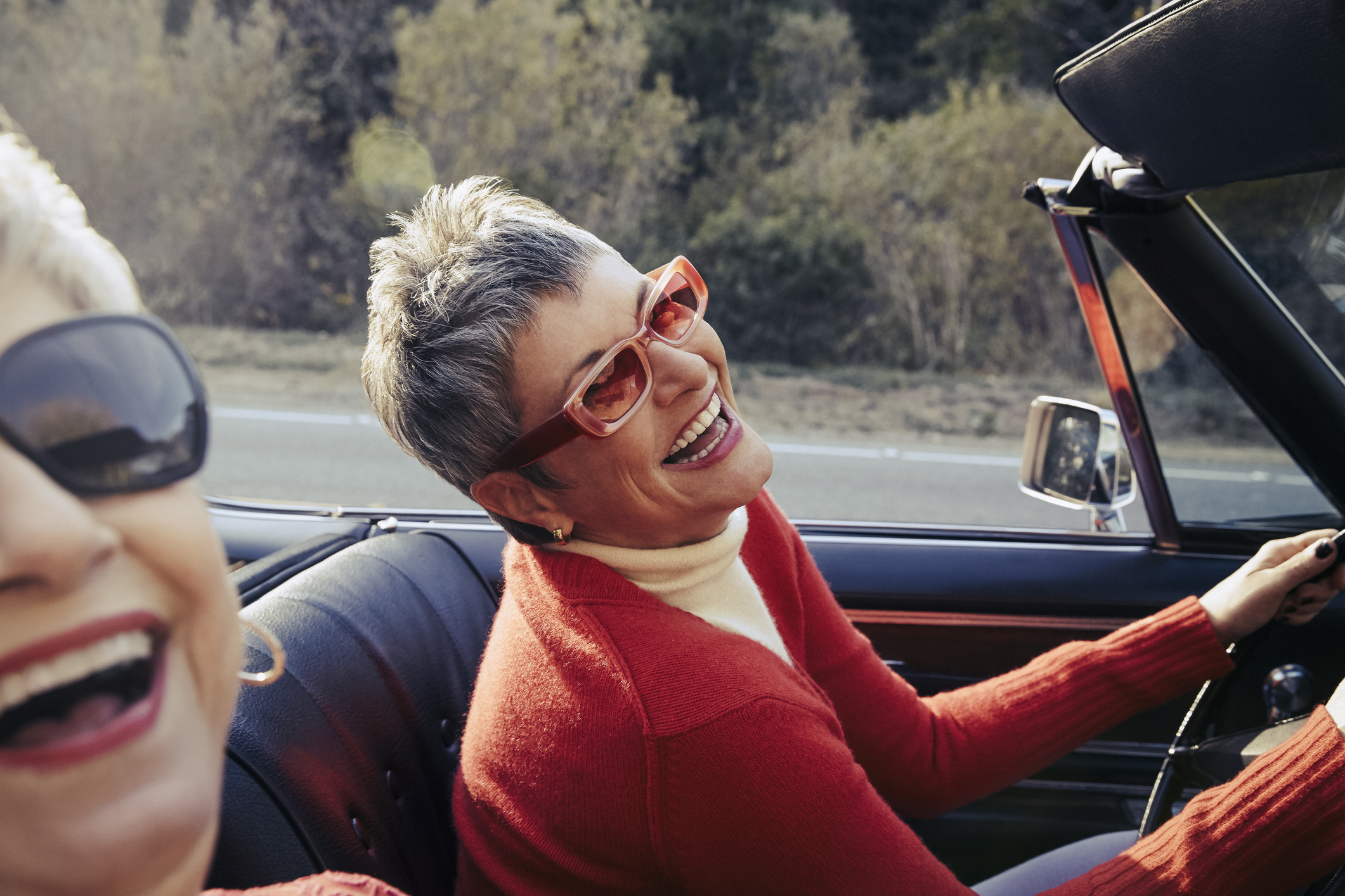 Two joyful women in a convertible, one driving and smiling broadly at the passenger