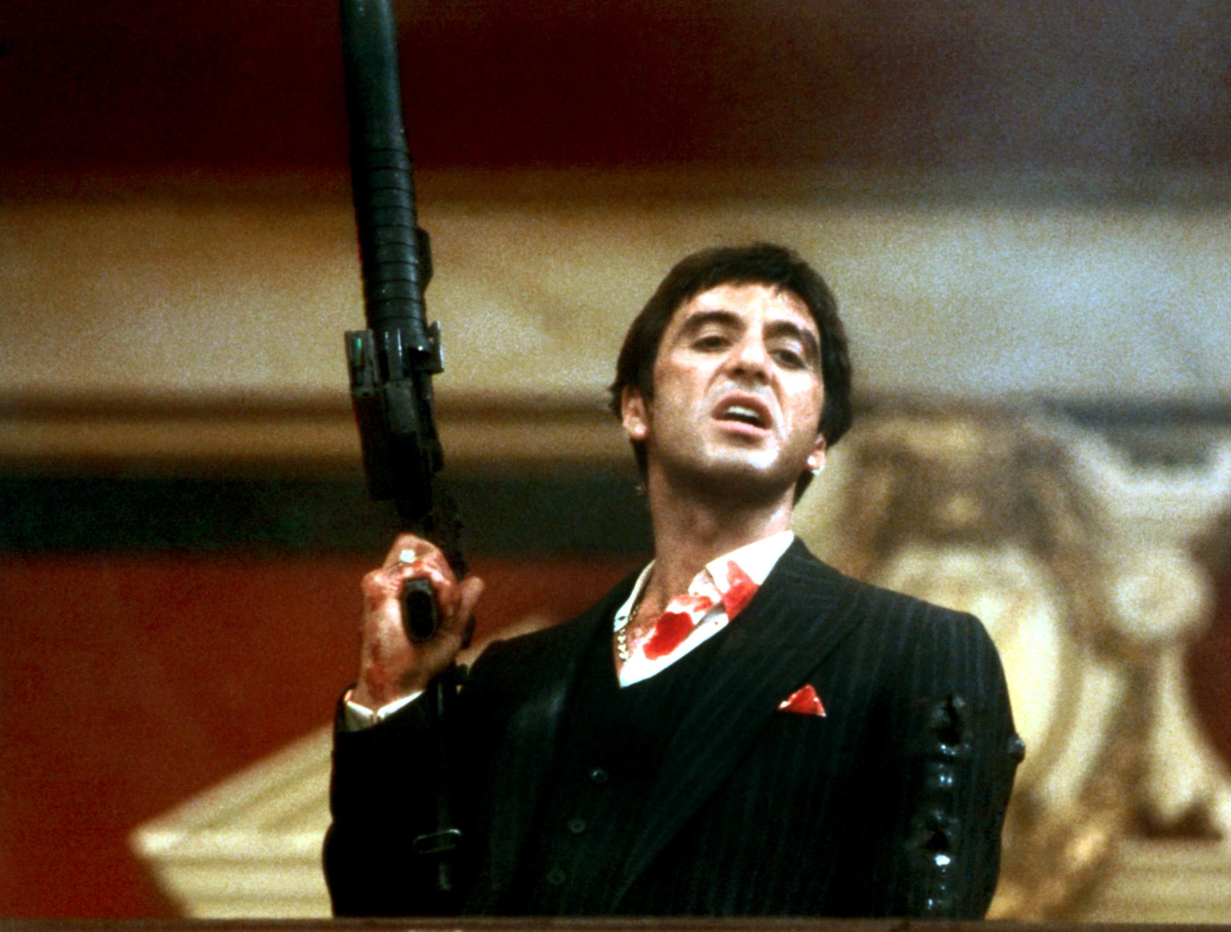 Al Pacino in Scarface, wearing a suit holding a gun with a bloodied face in a movie scene