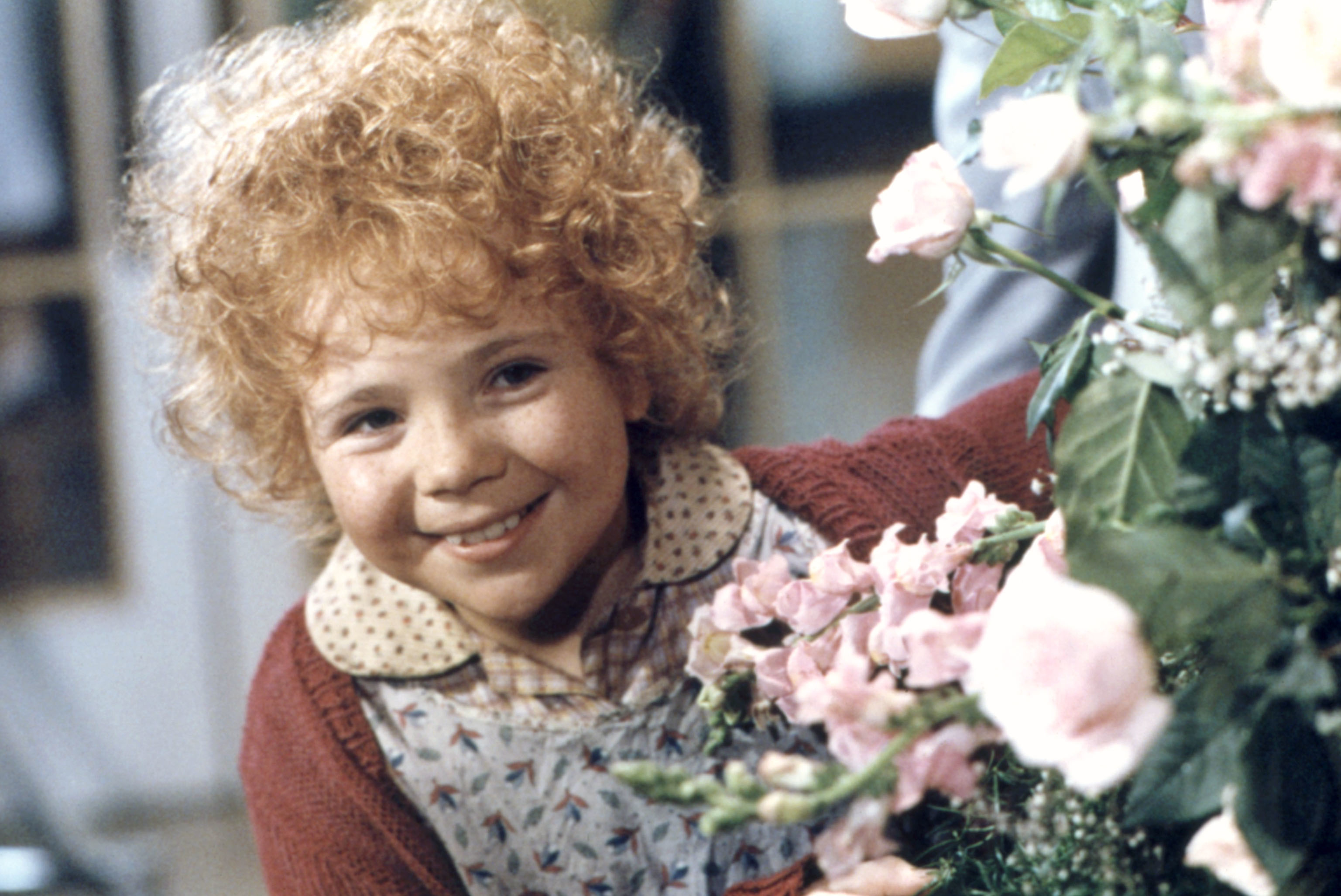 Annie from the musical &quot;Annie&quot; smiles, wearing a patterned dress and cardigan, holding a bouquet