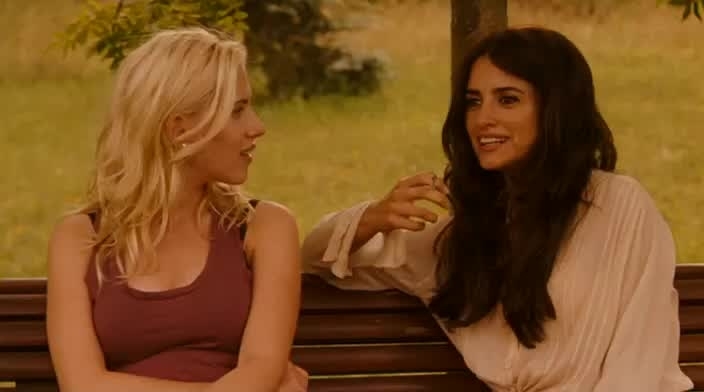 Two women sitting on a bench engaged in conversation, one in a sleeveless top, the other in a blouse