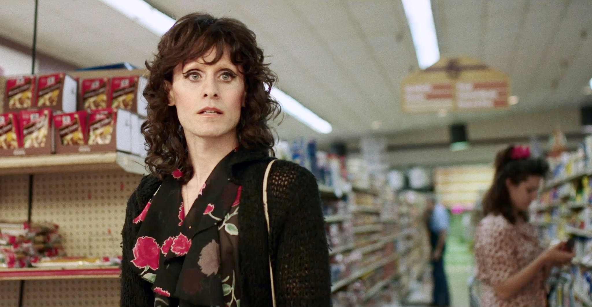 Woman in floral dress and cardigan stands worried in a supermarket aisle; another person in the background