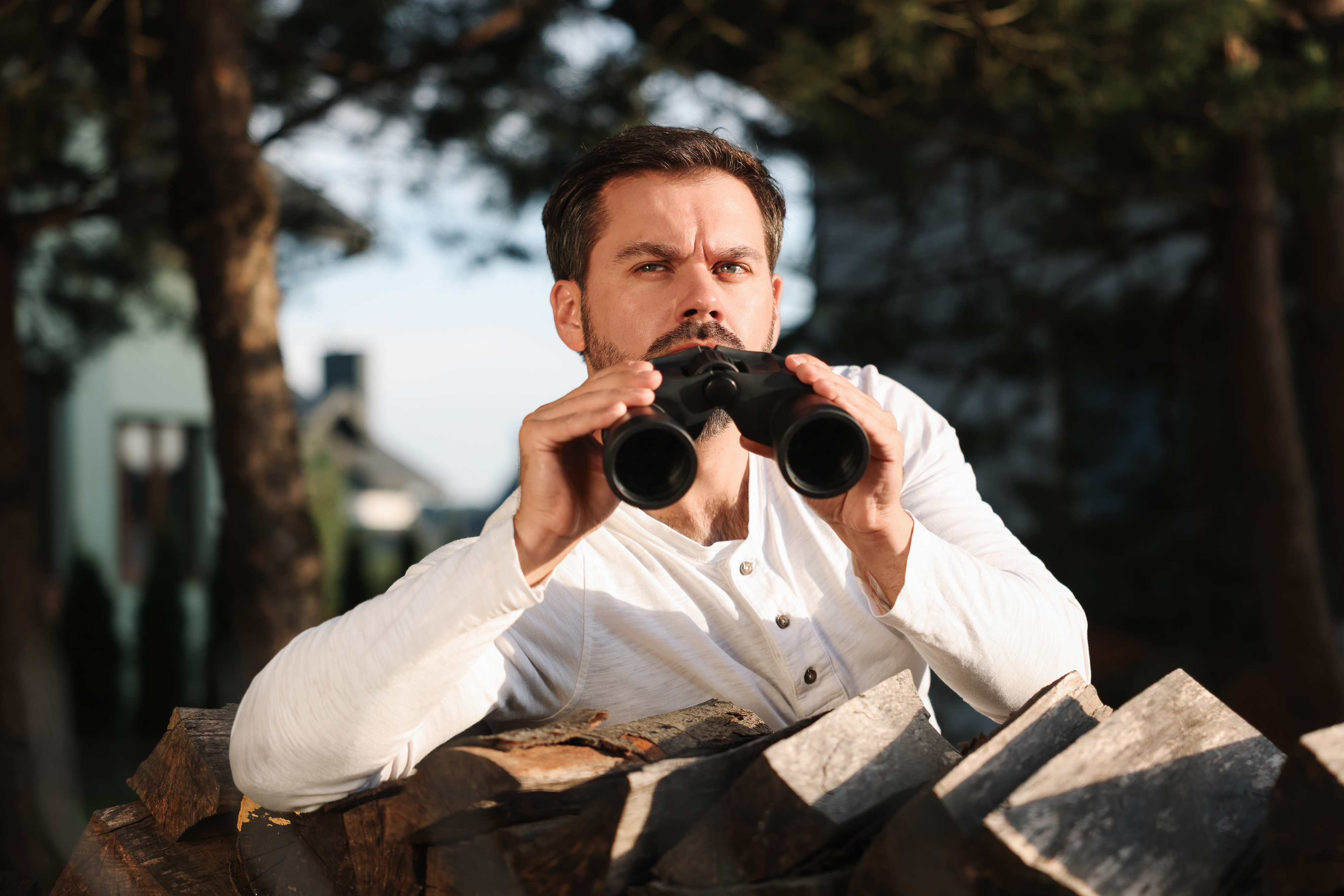 Man peering through binoculars over a pile of wood; expression focused