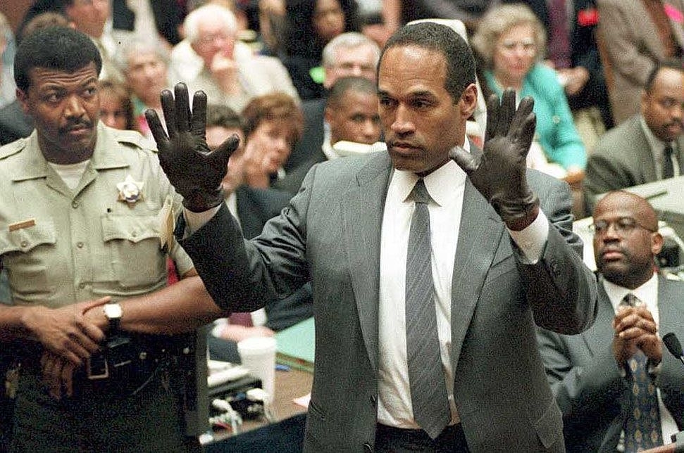 OJ in a courtroom; wears a suit, trying on gloves as others observe
