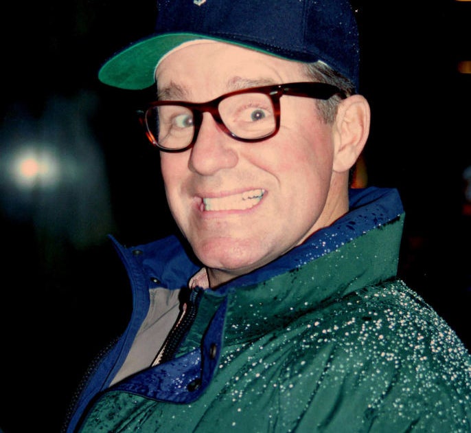 Phil Hartman in glasses making a surprised expression wears a cap and a jacket with raindrops
